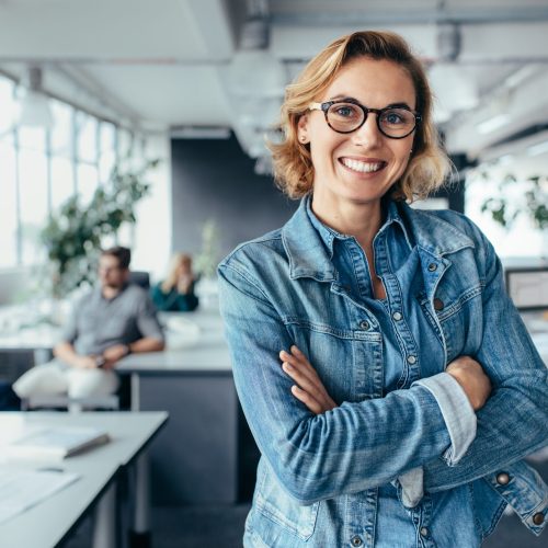 Successful creative female standing in office with colleagues in background. Happy woman executive standing in office with arms crossed.